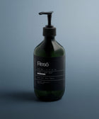 A dark green pump bottle labeled Hand & Body Wash, Ginger & Smoky Cardamom stands upright on a sleek blue-gray surface in soft light, showcasing a minimalist design and natural cleansers for an invigorating wash.