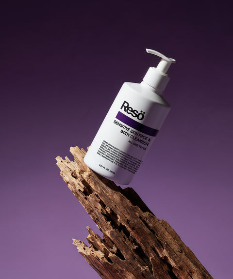 A white RESÖ bottle labeled Sensitive Skin Face & Body Cleanser balances on textured wood, set against a purple gradient background.