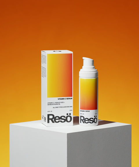 A bottle and box of Vitamin C Serum sit on a white pedestal with an orange gradient backdrop. The packaging features a white base and yellow-to-orange gradient label, emphasizing this serum’s antioxidant and brightening skincare benefits.