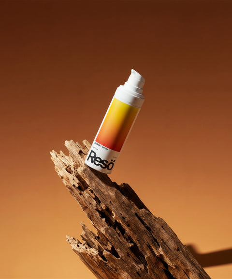 A cylindrical Vitamin C Serum bottle with a gradient yellow-orange label and white pump cap stands upright on textured, broken wood against a warm brown background.
