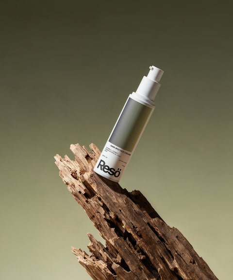 A bottle of RESÖ Dark Spot Face Cream with Kojic Acid and a white pump dispenser stands upright on textured, weathered wood against a gradient olive green background—ideal for targeting dark spots.