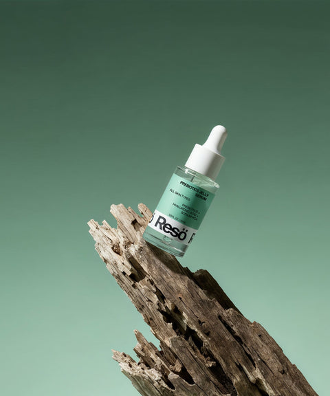 A dropper bottle of RESÖ Bioactive Prebiotics Jelly Serum, designed for soothing hydration, is balanced on a piece of weathered driftwood against a gradient green background.