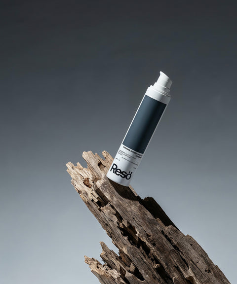 A white and gray bottle of RESÖ Ceramide Barrier Night Cream with ceramides and hyaluronic acid stands upright on rugged driftwood against a gradient gray background.