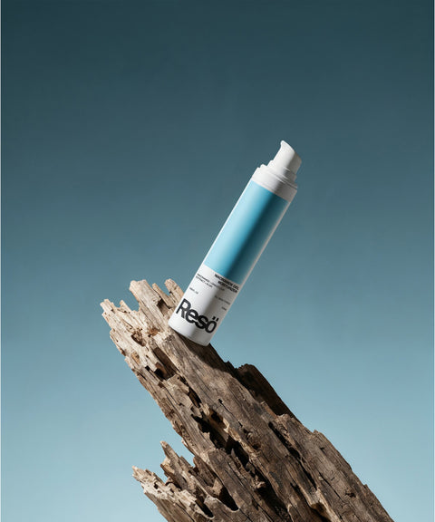 A white and blue cylindrical bottle of RESÖ Niacinamide Gel Moisturiser is balanced on rugged driftwood, set against a gradient blue background.
