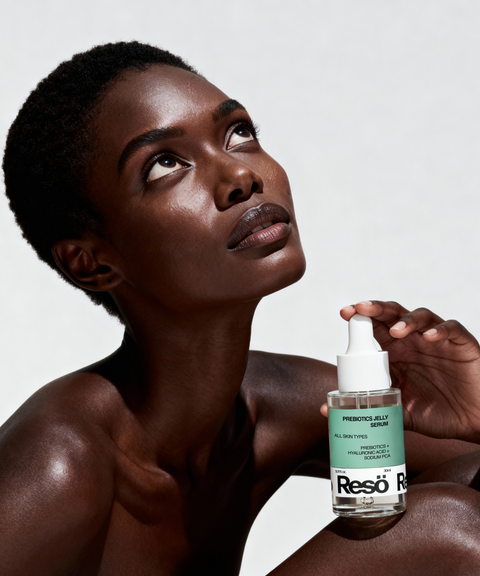 A woman with short hair looks upward while holding a bottle of Resö Prebiotics Jelly Serum in her hand against a plain background.