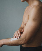 A shirtless person applies Silk Skin Body Serum, Woody Amber & Leather, to their forearm against a plain gray background. Only their upper body and arm are visible, emphasizing the elegance of luxury body care.