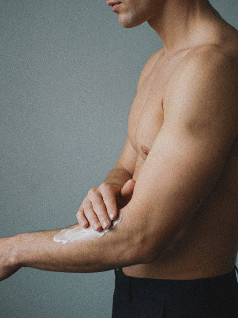 A shirtless person applies Silk Skin Body Serum, Woody Amber & Leather, to their forearm against a plain gray background. Only their upper body and arm are visible, emphasizing the elegance of luxury body care.
