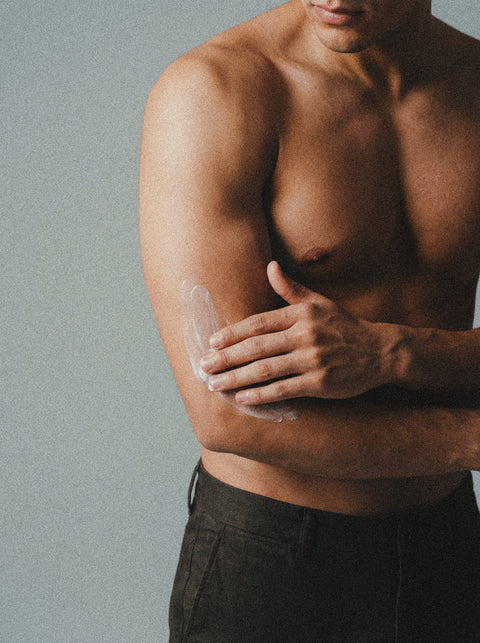 A shirtless person applies Silk Skin Body Serum, Spices & Sandalwood, to their upper arm with one hand against a plain neutral background. Visible are the torso, arms, and part of the lower face.