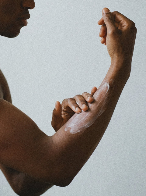 A person is applying lotion to their forearm, gently rubbing it in with their fingers. The background is plain and light-colored, focusing attention on the arm and the action.