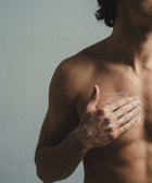 A shirtless person applies 5% Urea + 2% Panthenol Body Cream to their chest with their right hand against a neutral background, focusing on the upper torso and arm, suggesting skincare use for concerns like keratosis pilaris.