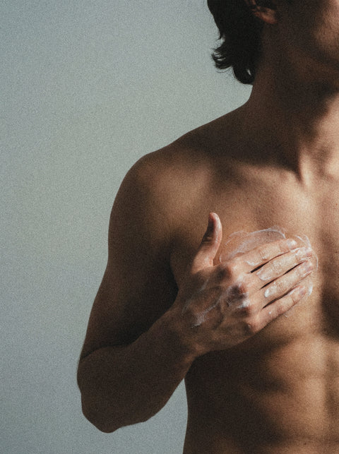 A shirtless person applies 5% Urea + 2% Panthenol Body Cream to their chest with their right hand against a neutral background, focusing on the upper torso and arm, suggesting skincare use for concerns like keratosis pilaris.