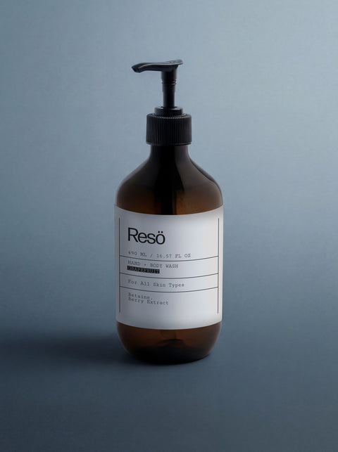 A brown pump bottle labeled Hand & Body Wash, Grapefruit stands upright on a plain, light gray surface with a gradient background. The label highlights hydrating botanic berry extract and natural surfactant for an effective citrus body wash.