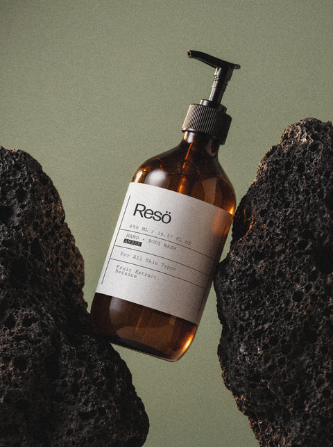 A brown pump bottle labeled "Hand & Body Wash, Patchouli & Amber Vanilla" is set between two black volcanic rocks against a muted green background.