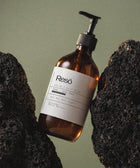 A brown glass pump bottle labeled Hand & Body Wash, Grapefruit with natural surfactant sits between black volcanic rocks on a muted green background. Suitable for all skin types, it contains betaine and persimmon extract for a refreshing citrus cleanse.