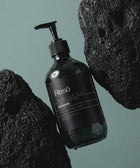 A dark-green pump bottle labeled Hand & Body Wash, Ginger & Smoky Cardamom rests between two textured black volcanic rocks on a matte green background. Water droplets and a minimalist black-and-white label accent the bottle.