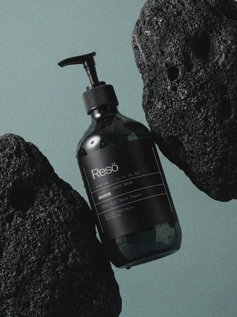 A black pump bottle labeled Hand & Body Wash, Peppermint & Dark Cedar sits on a greenish surface between large black rocks, water droplets visible on the bottle. The cleanser offers a nourishing peppermint and cedar scent.