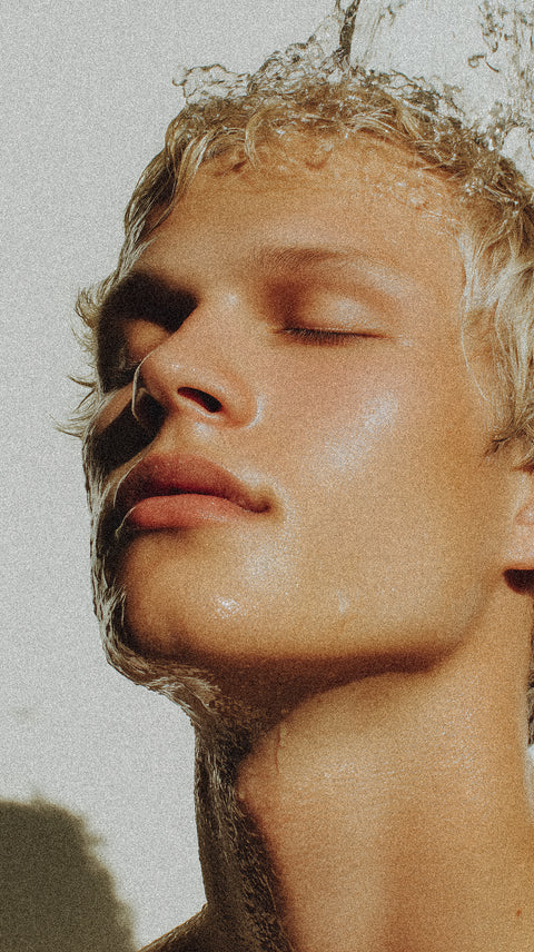 Close-up of a young person with short blond hair and closed eyes, tilting their head back as water splashes over their face, highlighting smooth skin and a peaceful expression.