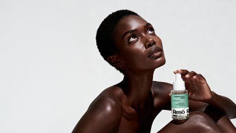 A woman with short hair and glowing skin looks upward while holding a clear bottle of skincare serum labeled Resō, set against a plain white background.