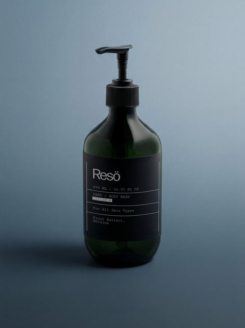 A dark green pump bottle labeled Hand & Body Wash, Ginger & Smoky Cardamom stands upright on a sleek blue-gray surface in soft light, showcasing a minimalist design and natural cleansers for an invigorating wash.