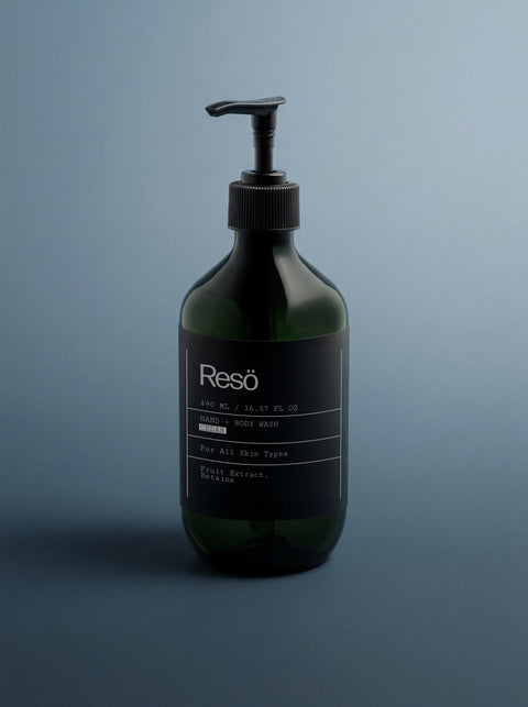 A dark green pump bottle labeled Hand & Body Wash, Peppermint & Dark Cedar sits on a blue-gray background. The minimalist label shows 490 ml and notes a nourishing formula for all skin types with fruit extract and a peppermint cedar scent.