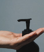 A hand is held open beneath the pump of a black bottle of Silk Skin Body Serum, Woody Amber & Leather, ready to dispense hydrating serum against a plain gray background.