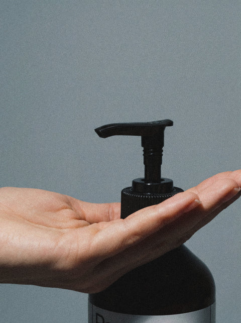 A hand is held open beneath the pump of a black bottle of Silk Skin Body Serum, Woody Amber & Leather, ready to dispense hydrating serum against a plain gray background.