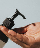 A close-up of a hand pressing the pump of a brown Silk Skin Body Serum, Spices & Sandalwood bottle, ready to dispense hydrating serum into an open palm, set against a neutral background.