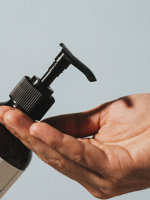 A close-up of a hand pressing the pump of a brown Silk Skin Body Serum, Spices & Sandalwood bottle, ready to dispense hydrating serum into an open palm, set against a neutral background.