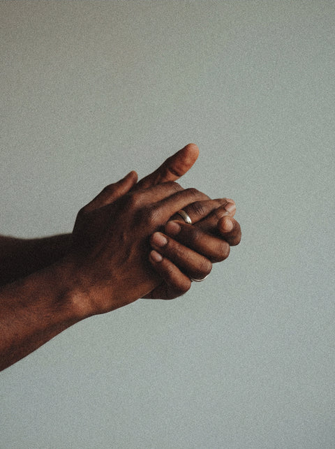 A close-up of two hands clasped together, one adorned with a simple ring, bathed in soft light that highlights the nourished skin—showcasing the gentle care of Silk Skin Hand Cream, Woody Amber & Leather.
