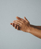 Two hands are held together against a light gray background. One, wearing a simple silver ring, gently touches the other—showcasing soft, calm skin after using Silk Skin Hand Cream, Spices & Sandalwood.