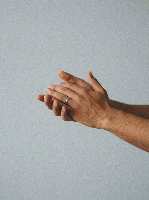 Two hands are held together against a light gray background. One, wearing a simple silver ring, gently touches the other—showcasing soft, calm skin after using Silk Skin Hand Cream, Spices & Sandalwood.