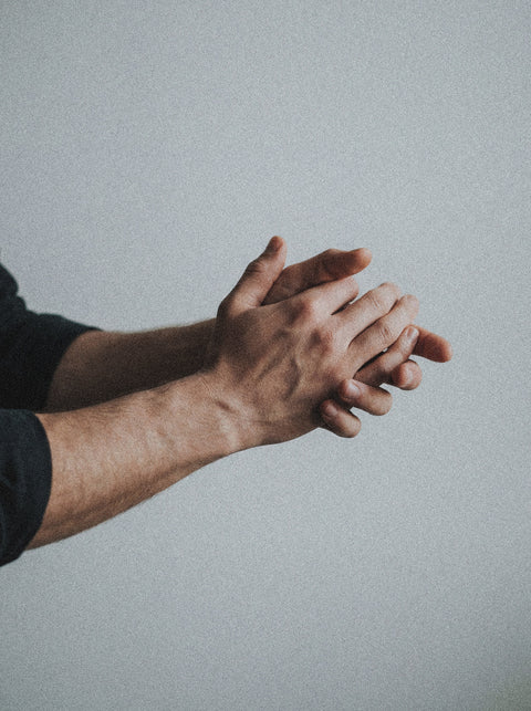 A close-up of a person’s hands clasped together against a plain, light gray background. The person is wearing a dark long-sleeved shirt.