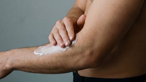 A person applies a white cream or lotion to their forearm, using their right hand. The person is shirtless and the background is plain and neutral.