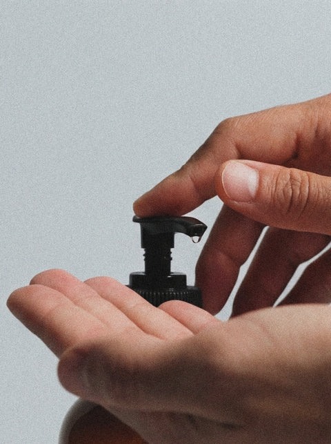 A close-up shows someone pressing a pump bottle of Hand & Body Wash, Peppermint & Dark Cedar, dispensing the wash onto their hand against a plain gray background.