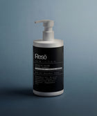 A white pump bottle of Keratin Volume Boost Shampoo sits on a gray surface with a gray-blue background. The black label highlights ingredients such as rosemary oil, keratin amino acids, and peppermint oil.