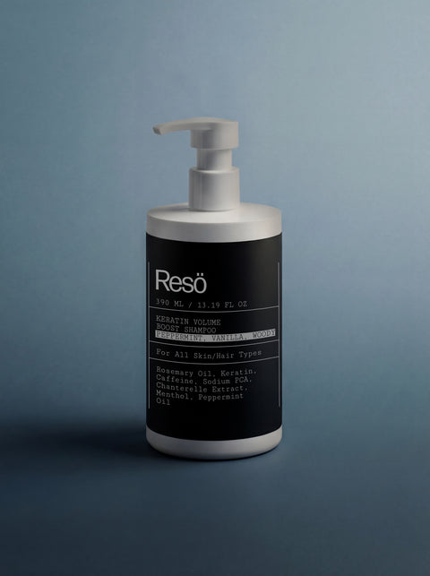 A white pump bottle of Keratin Volume Boost Shampoo sits on a gray surface with a gray-blue background. The black label highlights ingredients such as rosemary oil, keratin amino acids, and peppermint oil.