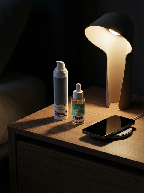 A bedside table with a modern lamp, a bottle of Resö skincare serum, a Resö pump bottle, and a smartphone resting on a wireless charger, all softly lit by the lamp.