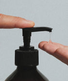 A close-up of two hands using a Hand & Body Wash, Patchouli & Amber Vanilla pump bottle, with a drop of clear liquid dispenser falling onto a fingertip against a plain light gray background.