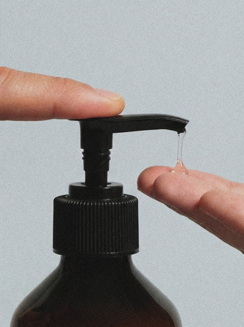 A close-up of two hands using a Hand & Body Wash, Patchouli & Amber Vanilla pump bottle, with a drop of clear liquid dispenser falling onto a fingertip against a plain light gray background.