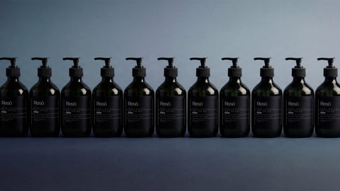 Twelve dark pump bottles labeled “Resó” are lined up in a row against a gradient gray-blue background, creating a minimalist and symmetrical display.