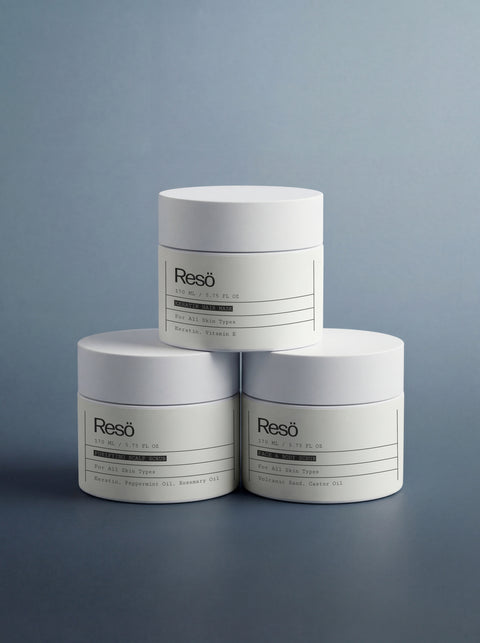 Three white cylindrical containers labeled SCRUB & MASK BUNDLE are stacked in a pyramid against a soft blue-gray background. Minimalist labels highlight active ingredients and note suitability for all skin types.