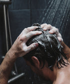 A person stands under a shower, washing their hair with Keratin Volume Boost Shampoo, lathering it into their wet, dark hair using both hands as water streams down from the showerhead above.