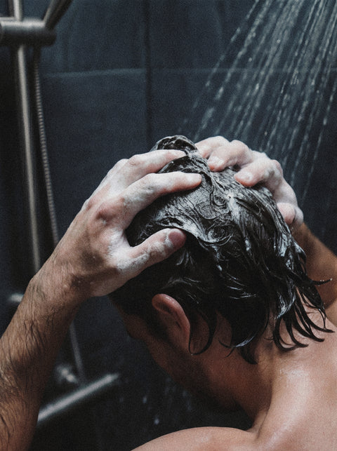 A person stands under a shower, washing their hair with Keratin Volume Boost Shampoo, lathering it into their wet, dark hair using both hands as water streams down from the showerhead above.