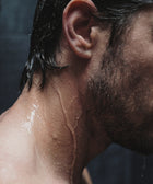 Close-up of the side of a man's face, neck, and shoulder with wet hair and water droplets. Dim lighting highlights the effects of Keratin Volume Boost Shampoo, a sulfate-free, volumizing formula that adds lift and shine to his hair.