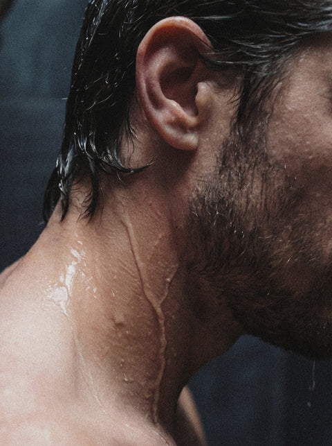 Close-up of the side of a man's face, neck, and shoulder with wet hair and water droplets. Dim lighting highlights the effects of Keratin Volume Boost Shampoo, a sulfate-free, volumizing formula that adds lift and shine to his hair.