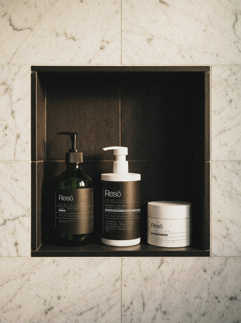 Three Resō branded bath products—liquid soap in a pump bottle, lotion in a larger pump bottle, and a round jar—are neatly arranged on a dark recessed shelf set into a marble-tiled wall.