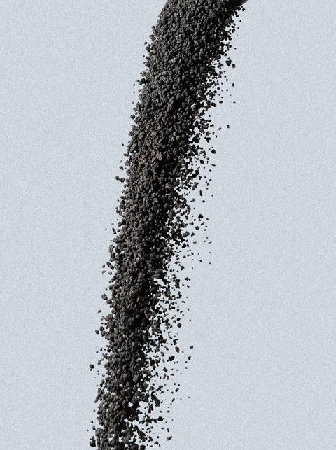 Granular particles of the Icelandic Volcano Face & Body Scrub are shown mid-air against a light gray background, forming a curved arc as the dark, volcanic ash-like bits scatter and fall.